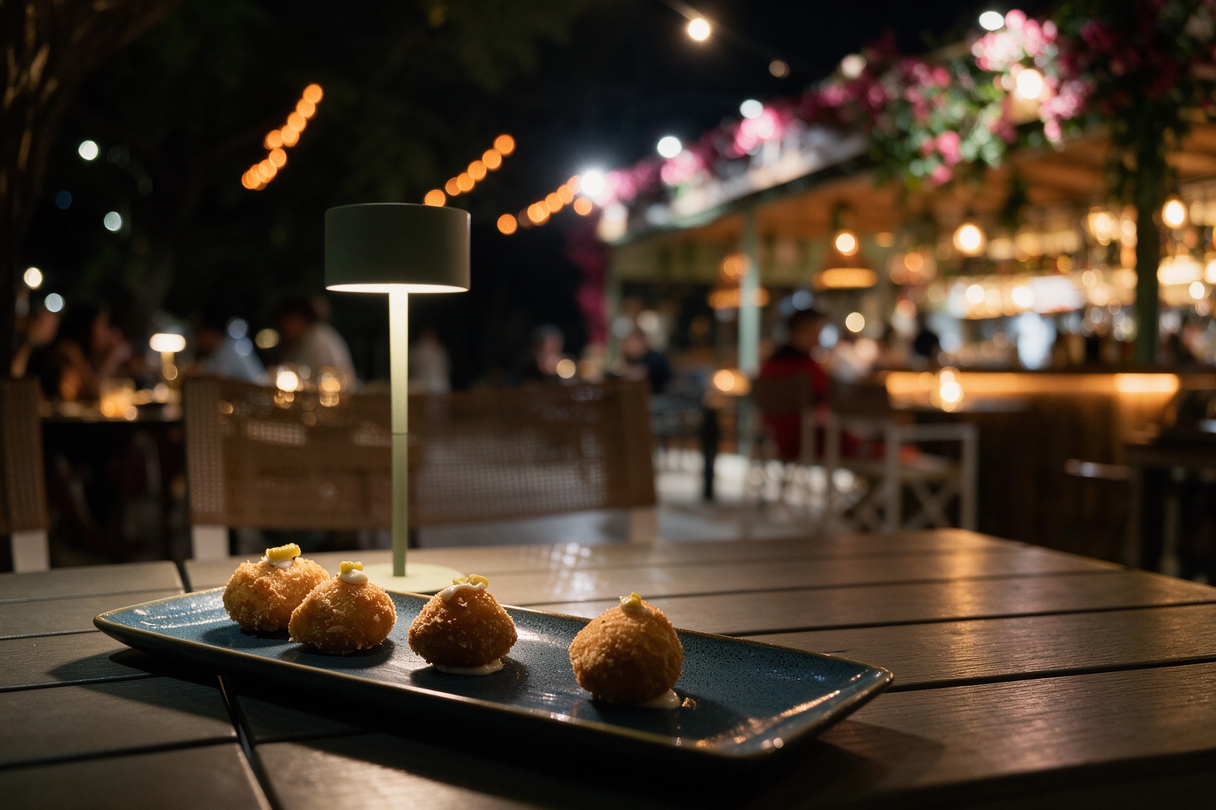 Terraza exterior romántica de noche con mesa de madera, luces de cadena y croquetas en pizarra